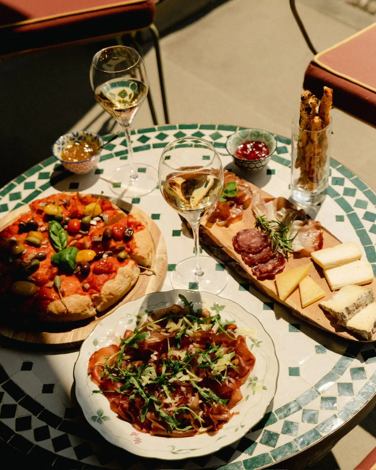 Sunlit mosaic table set with pizza, cured meats, cheese and glasses of Franciacorta